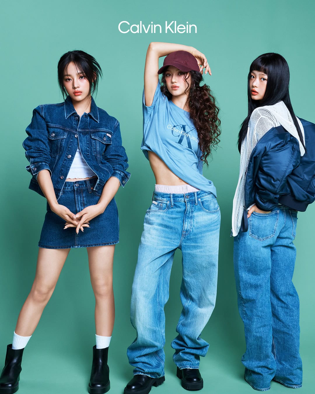 Three young female models in denim attire posing against a solid teal studio background for a Calvin Klein campaign.