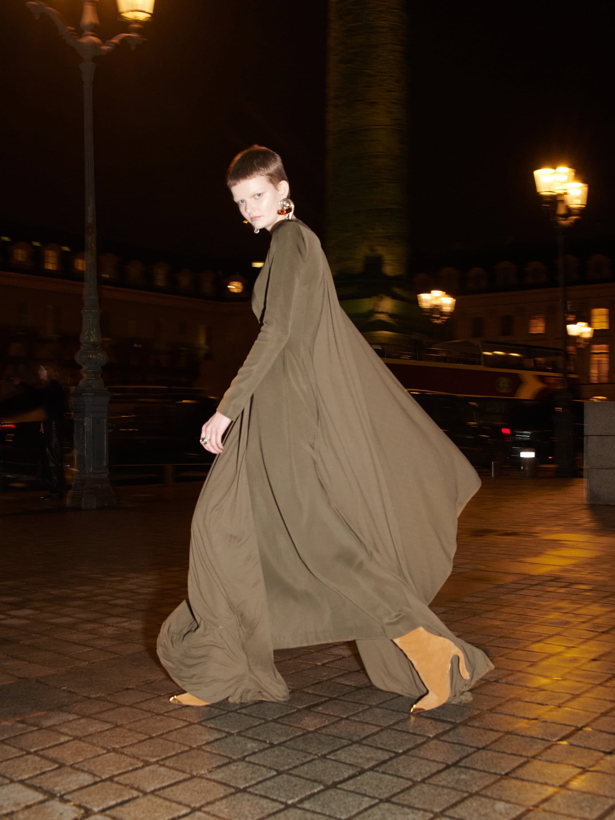 Model with short hair walking outside at night in a long olive green dress and heeled boots under street lamps