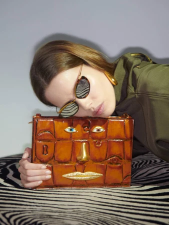 Model resting her head on a brown leather handbag with sculpted facial features wearing sunglasses