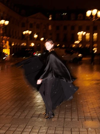 Model in black cape coat standing on a cobblestone square at night with blurred streetlights and glowing architecture in the background.