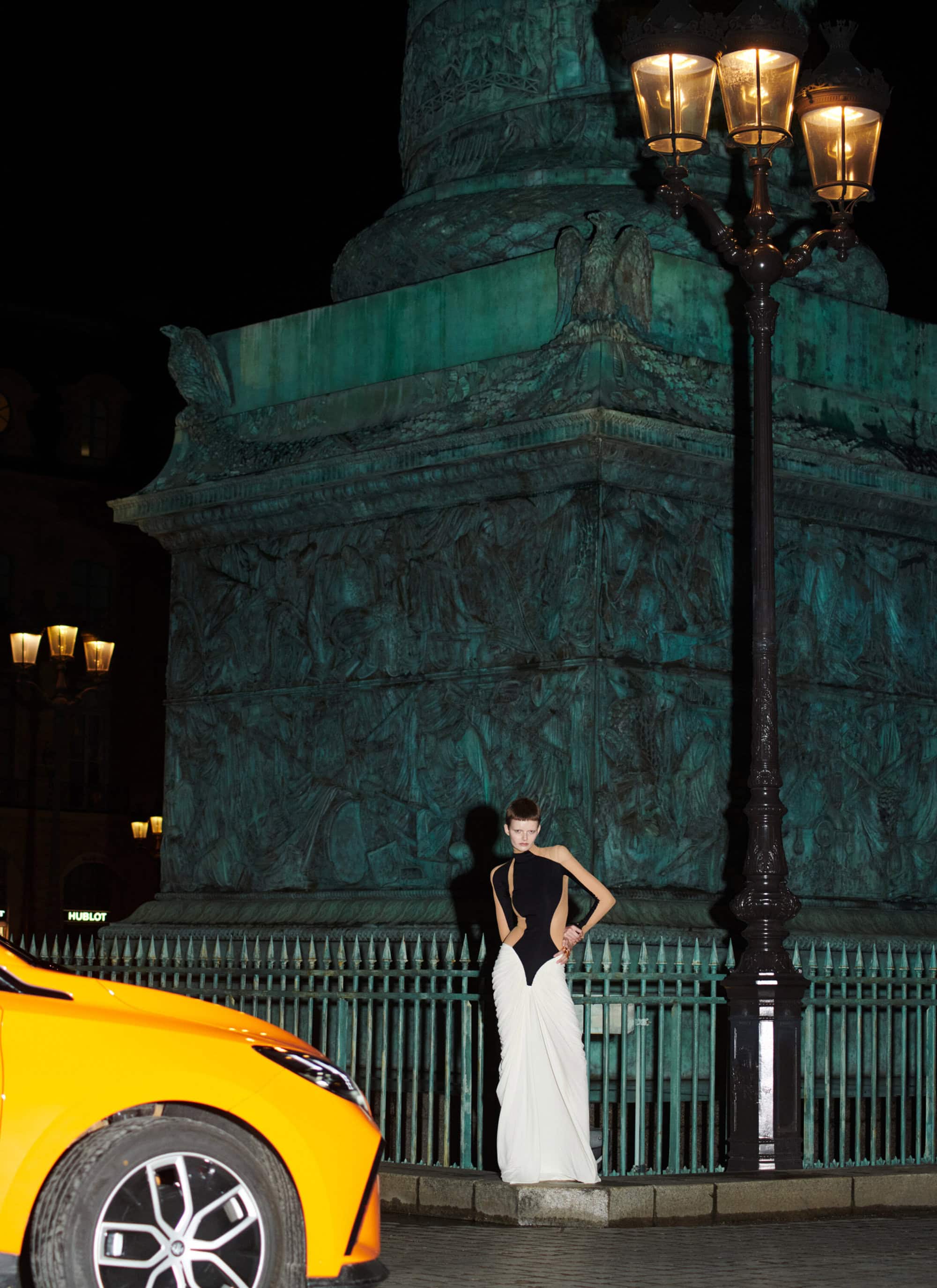 Model standing in front of Place Vendome column at night next to a lamppost and a yellow car for Schiaparelli