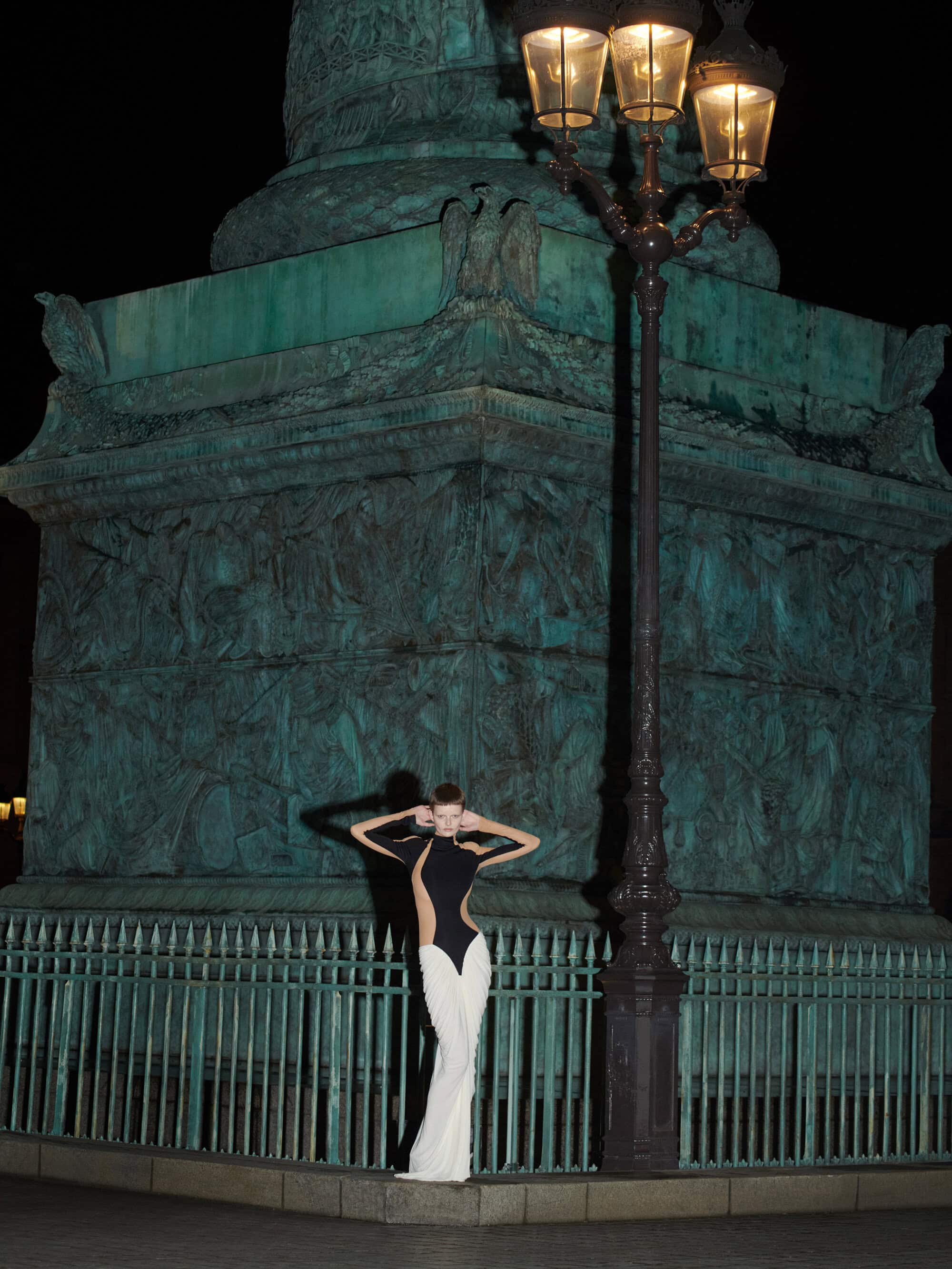 Model standing in front of a dark stone monument at night next to a tall ornate streetlamp for Schiaparelli Fall 2024 Ad Campaign