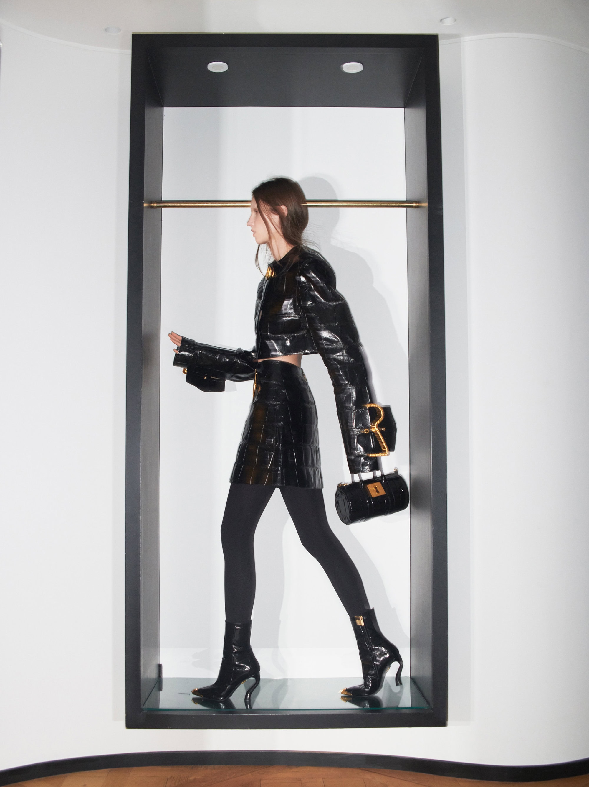 Model in black textured jacket and skirt holding a handbag inside a dark rectangular alcove