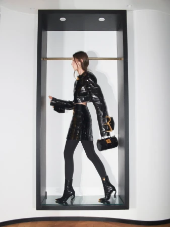 Model in black textured jacket and skirt holding a handbag inside a dark rectangular alcove