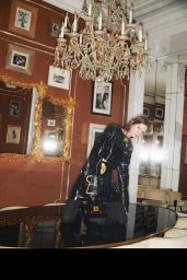 Model in shiny black vinyl jacket and skirt kneeling on a large oval table inside a room with terracotta walls and a crystal chandelier.