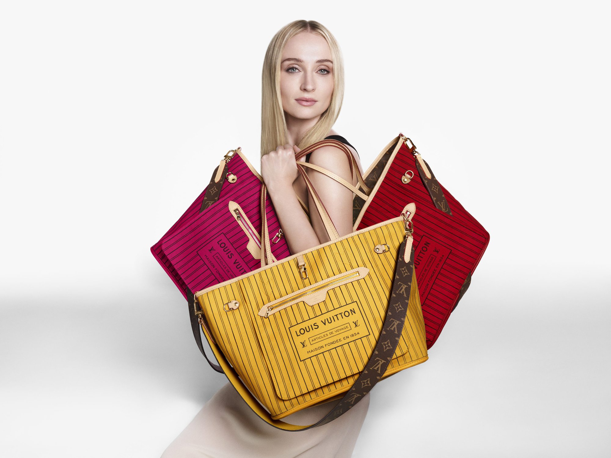 Model Sophie Turner holding multiple colorful Louis Vuitton Neverfull Inside Out handbags against a white studio background.