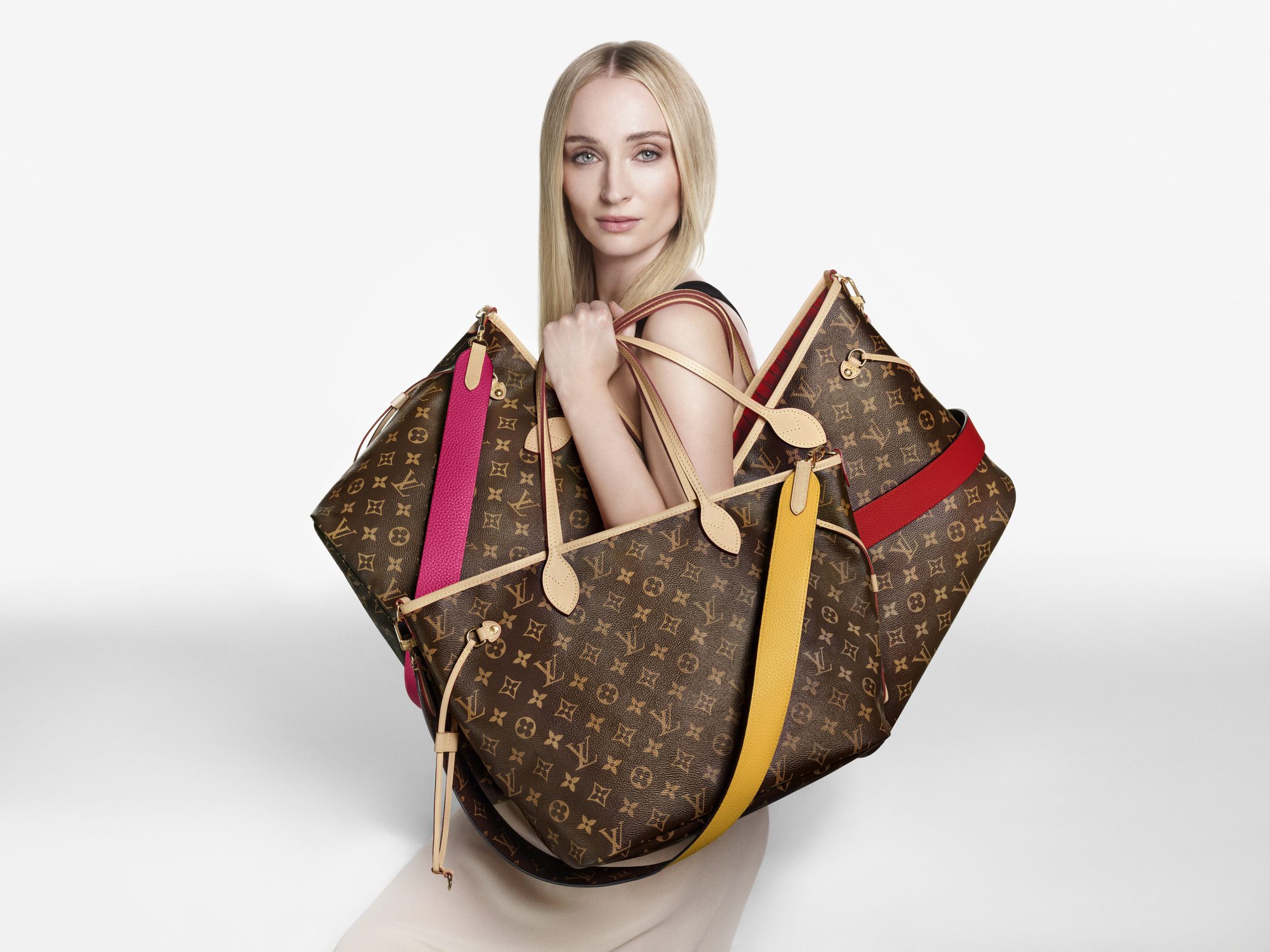 Sophie Turner holding multiple monogram canvas tote bags for the Louis Vuitton Neverfull Inside Out campaign against a white backdrop.