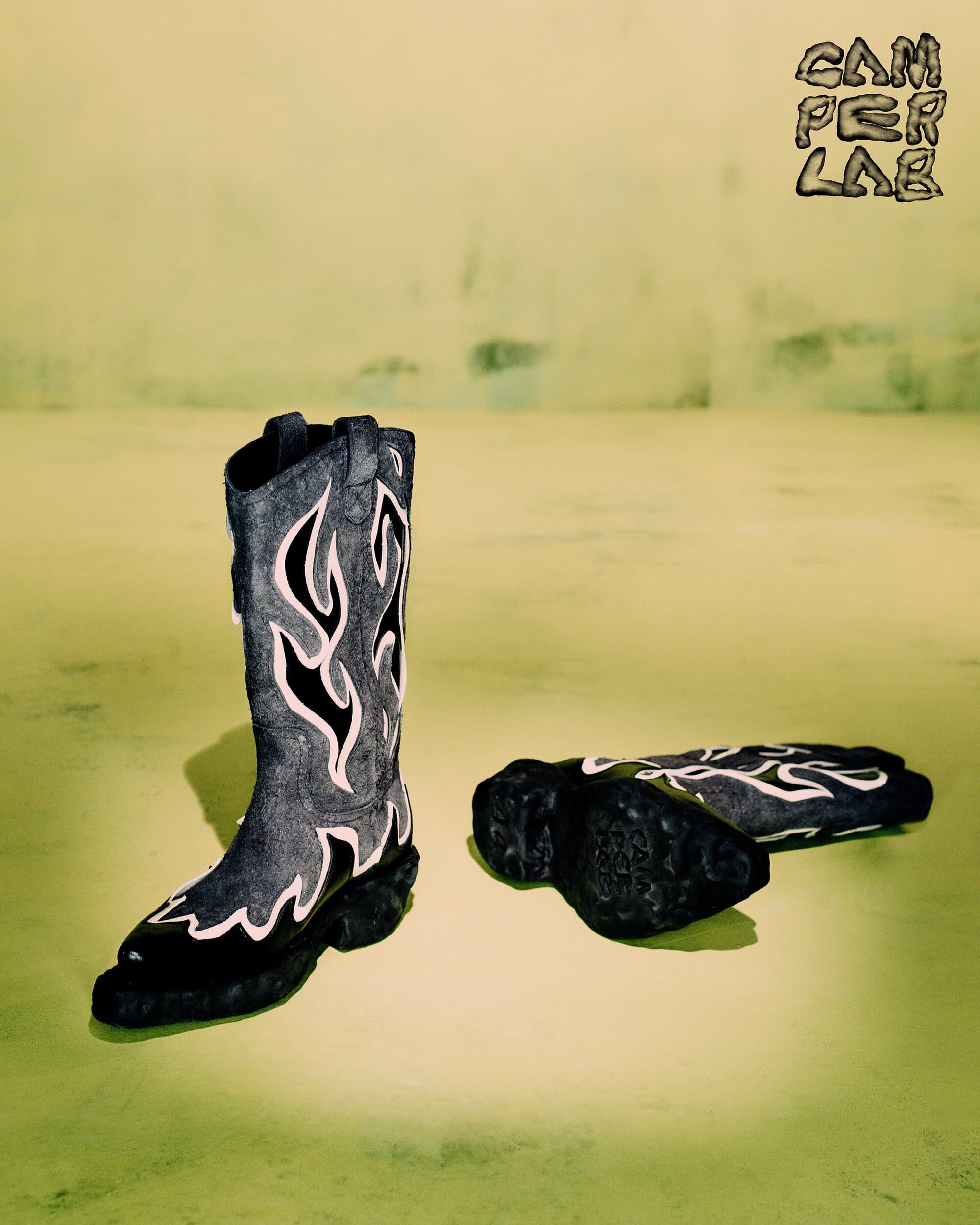 Black leather cowboy boots with white flame patterns displayed upright and lying down on a textured yellow-green studio background.