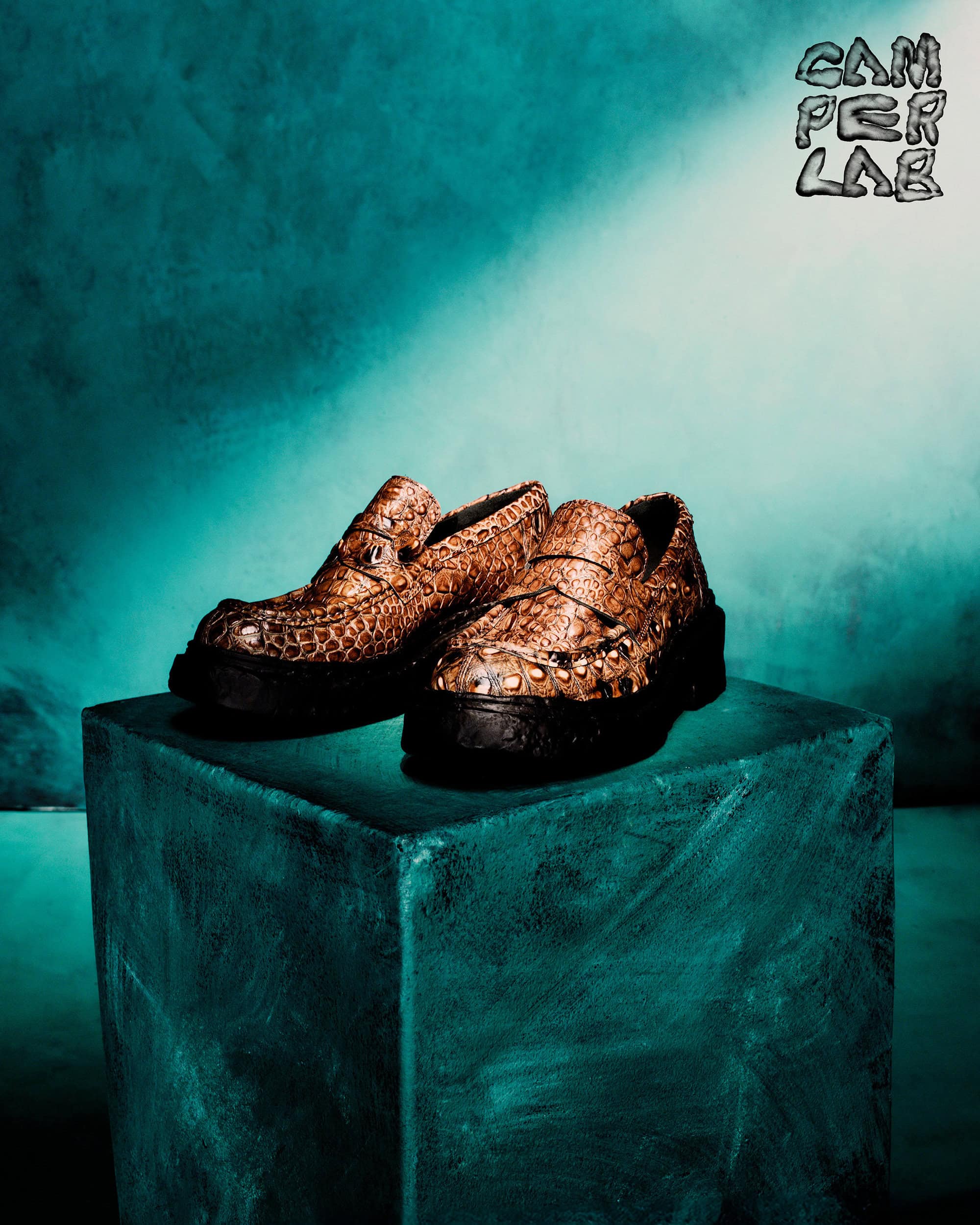 Brown embossed leather loafers with black platform soles resting on a teal textured block against a moody teal backdrop.
