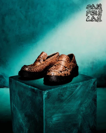 Brown embossed leather loafers with black platform soles resting on a teal textured block against a moody teal backdrop.