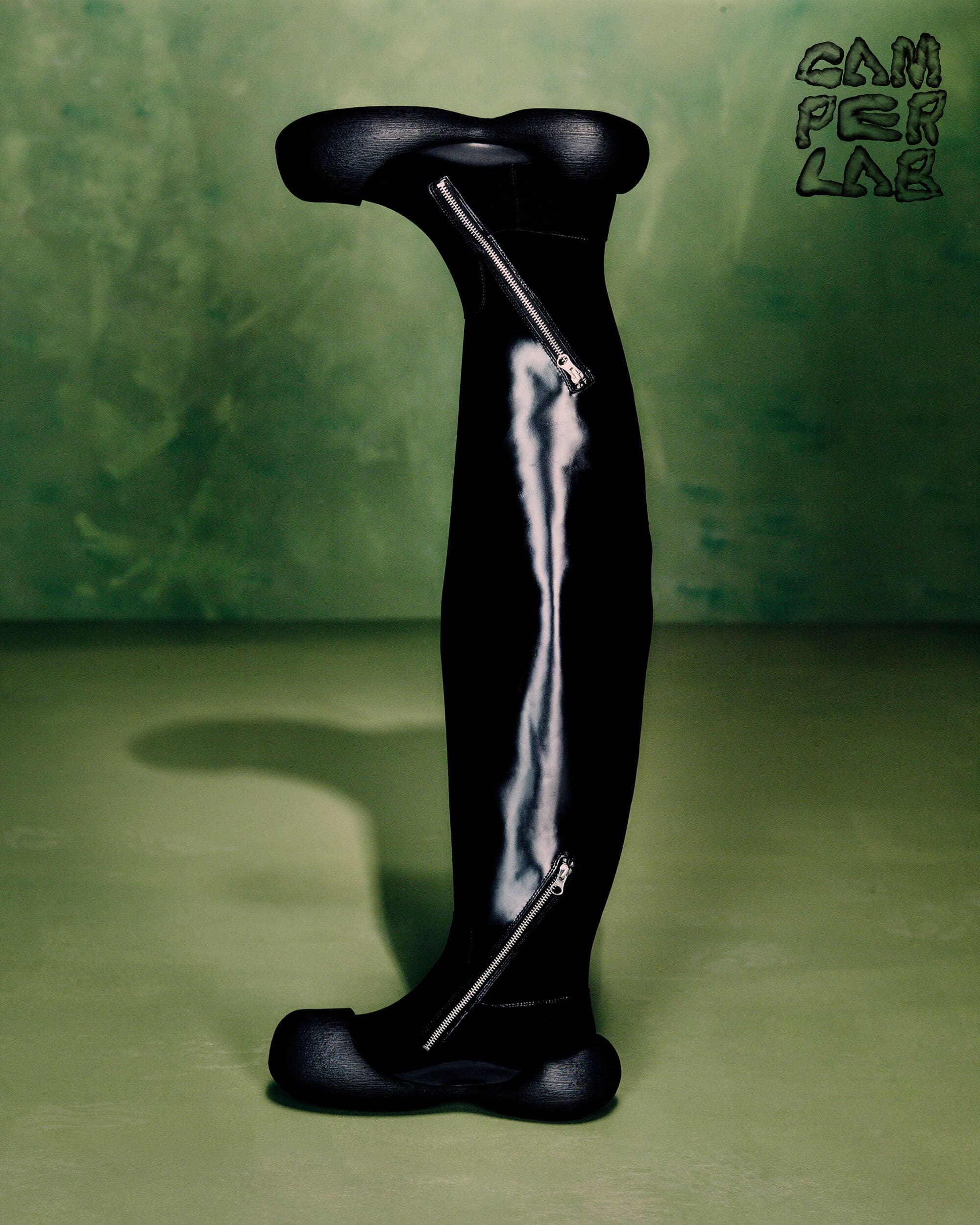Black tall boot with curved toe and heel and silver zippers against a mottled green studio background for Camperlab Fall 2024 campaign.