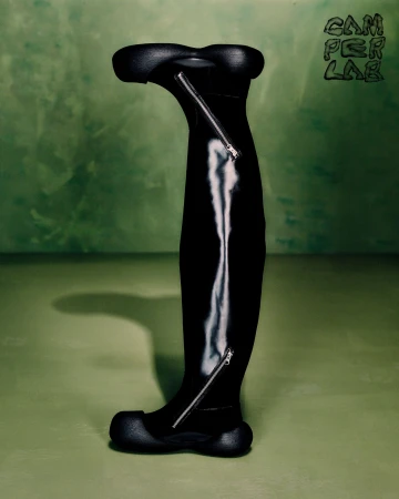 Black tall boot with curved toe and heel and silver zippers against a mottled green studio background for Camperlab Fall 2024 campaign.