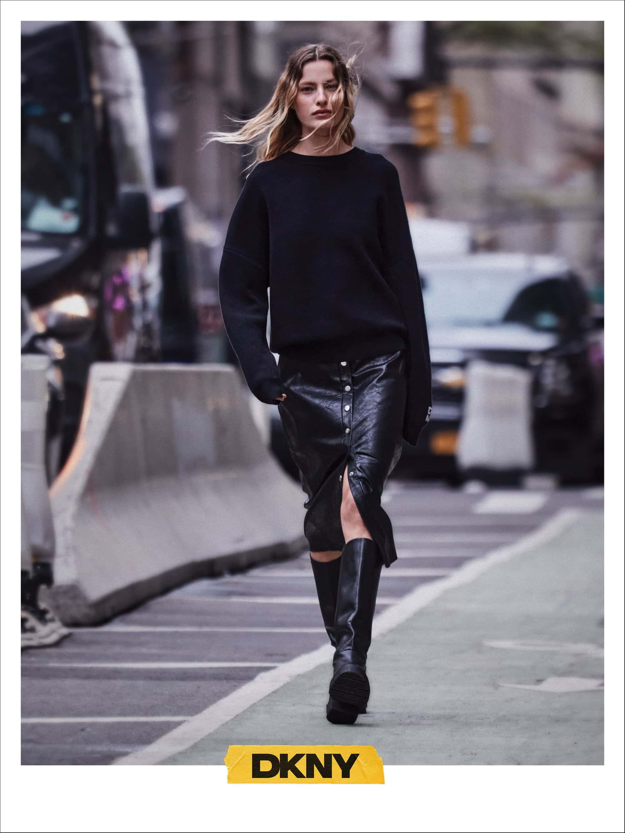 Model walking on a city street wearing a black sweater, black leather skirt, and tall boots for DKNY