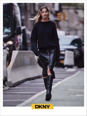 Model walking on a city street wearing a black sweater, black leather skirt, and tall boots for DKNY