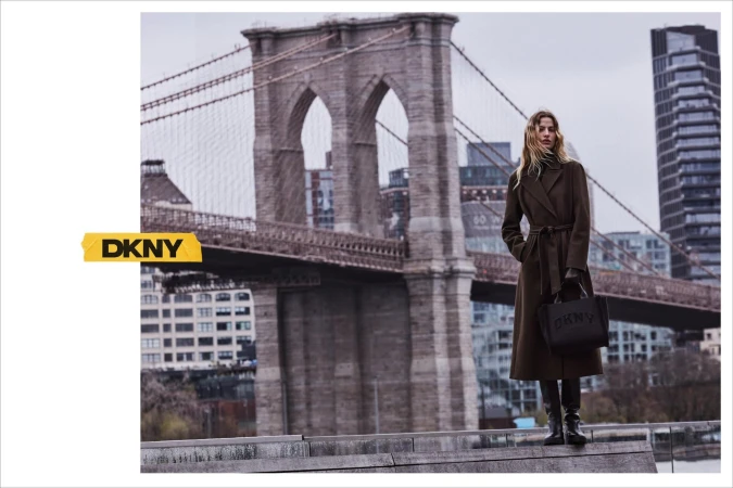 Model wearing a dark brown belted coat and boots holding a branded black handbag outdoors with the Brooklyn Bridge in the background.