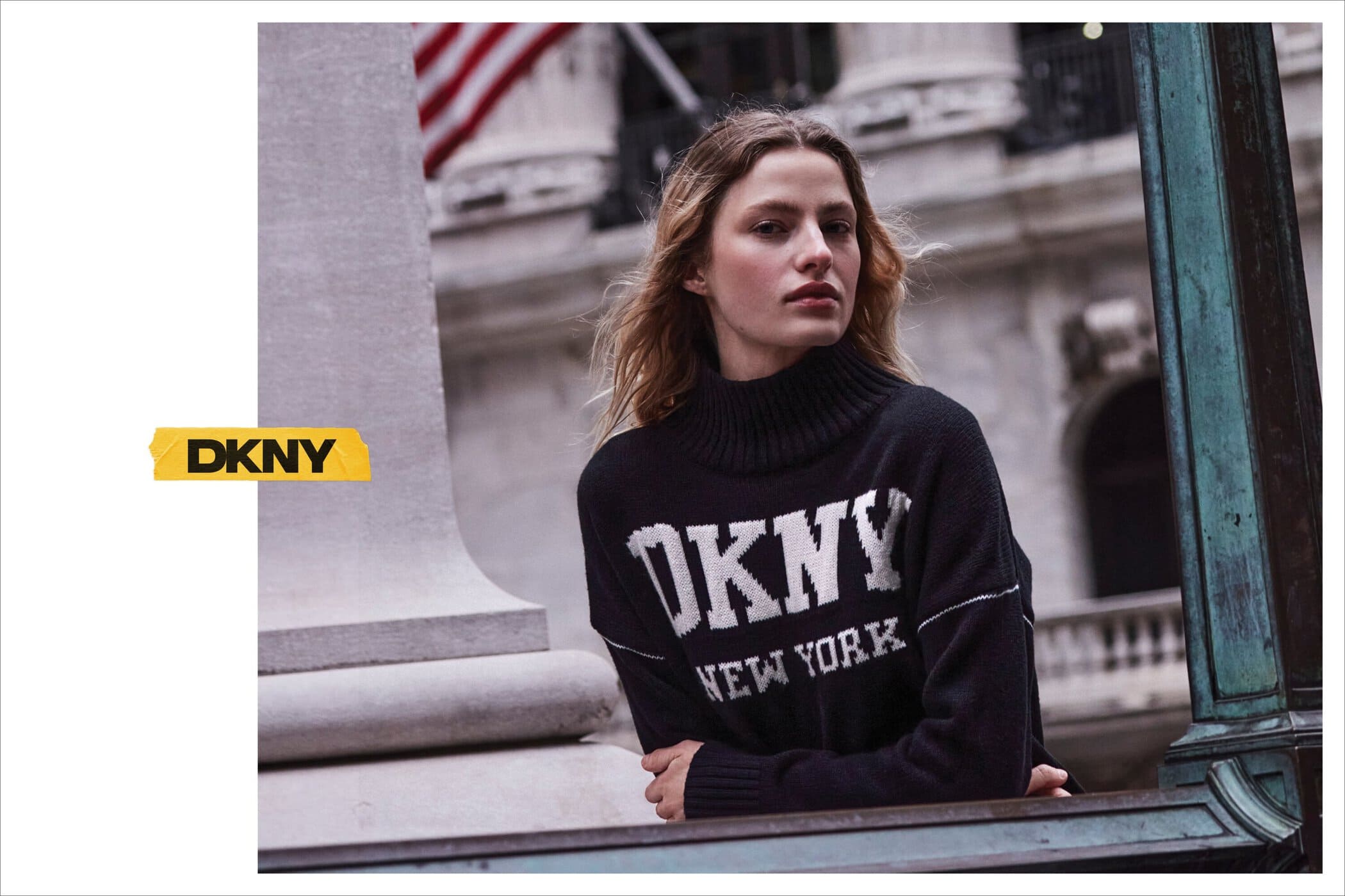 Model wearing a black turtleneck sweater with DKNY logo standing outdoors near classical stone architecture.