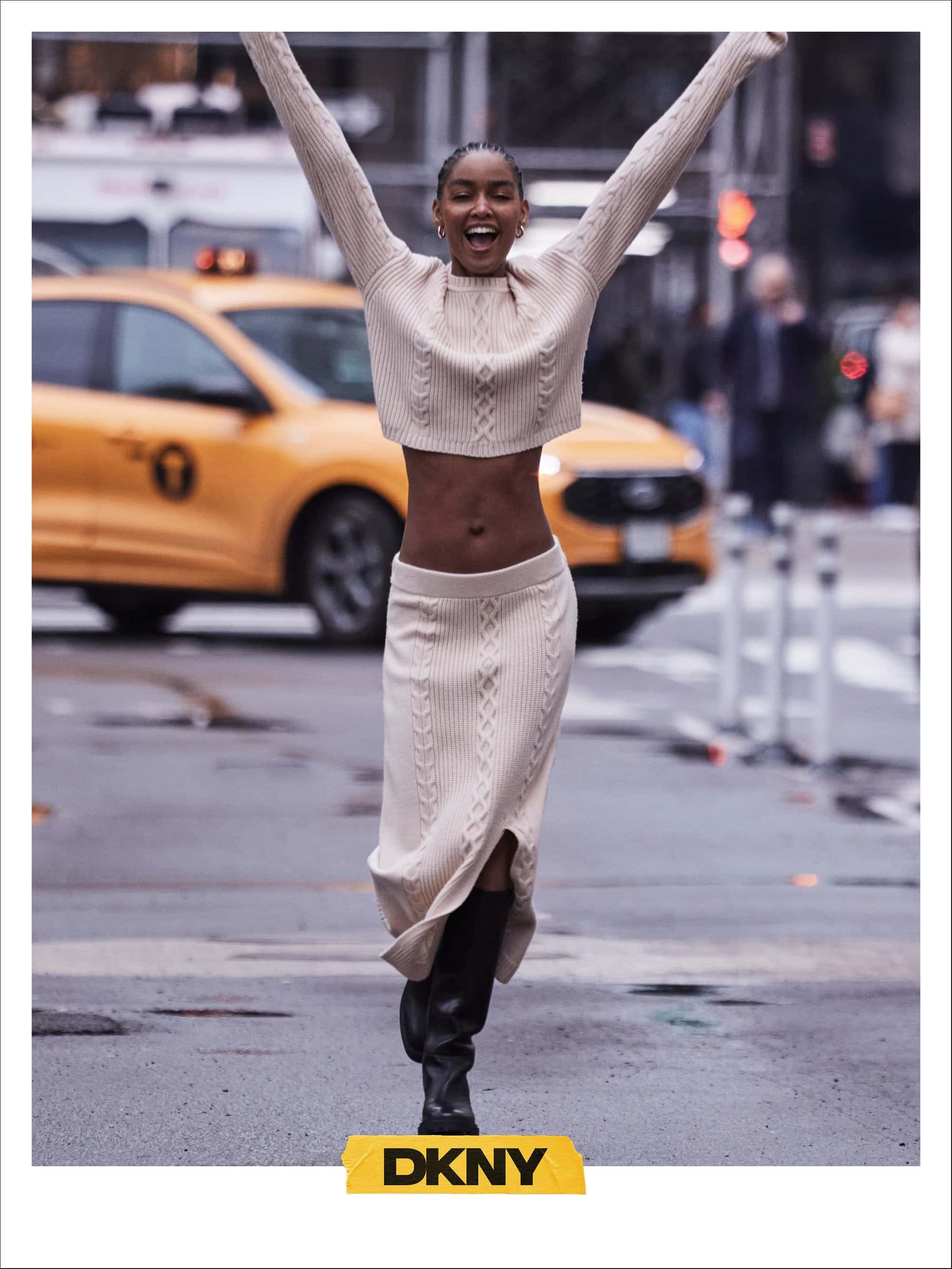 Model in cream knit sweater and skirt running on a city street with a yellow taxi in the background for DKNY Fall 2024 campaign