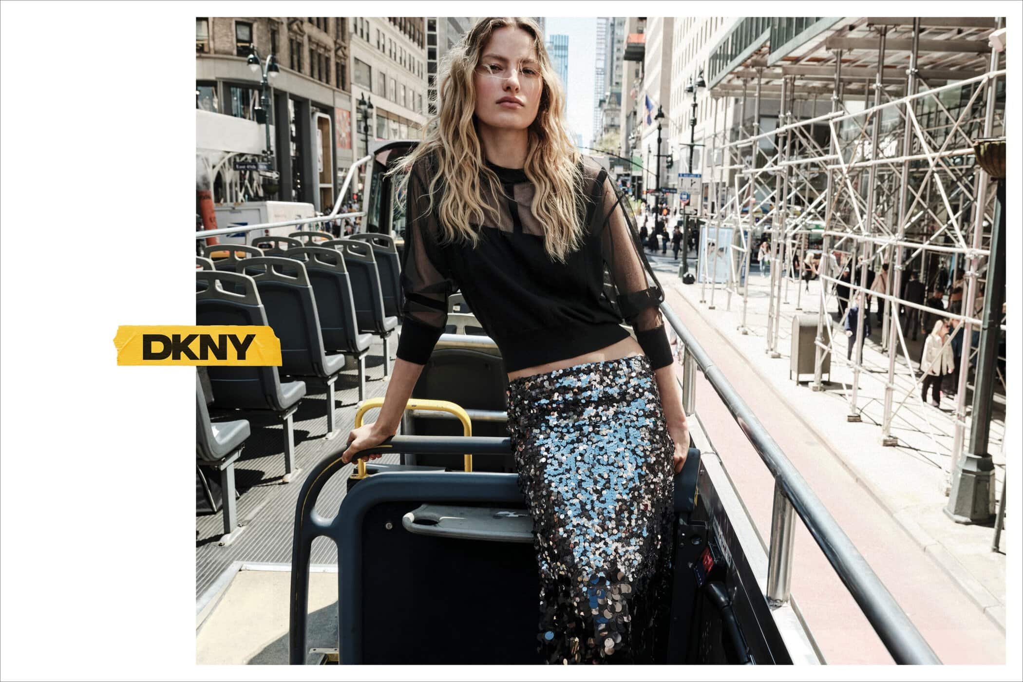 Model with blonde hair standing on an open top bus wearing a black sheer top and a sequin skirt in New York City