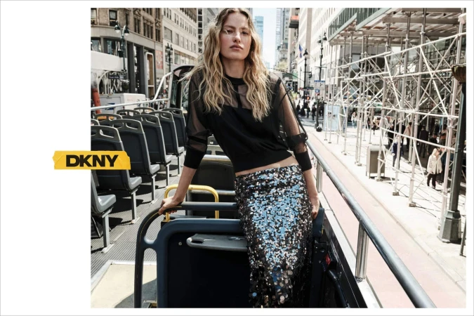 Model with blonde hair standing on an open top bus wearing a black sheer top and a sequin skirt in New York City