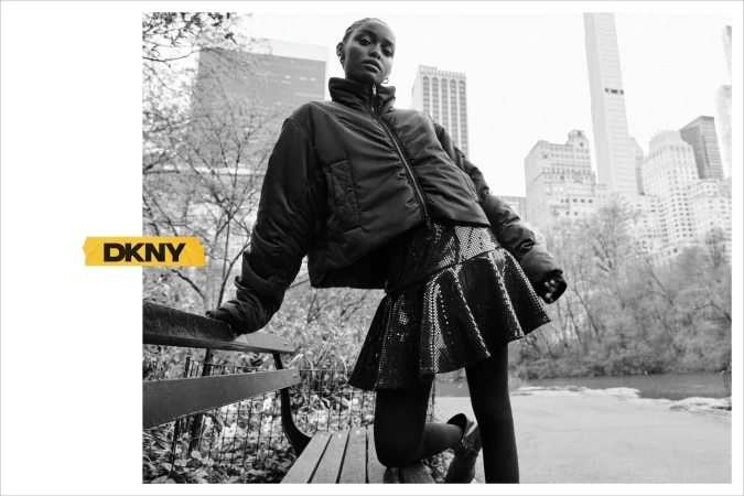 Black and white portrait of a model in a puffer jacket and sequined skirt posing by a park bench against a city skyline.