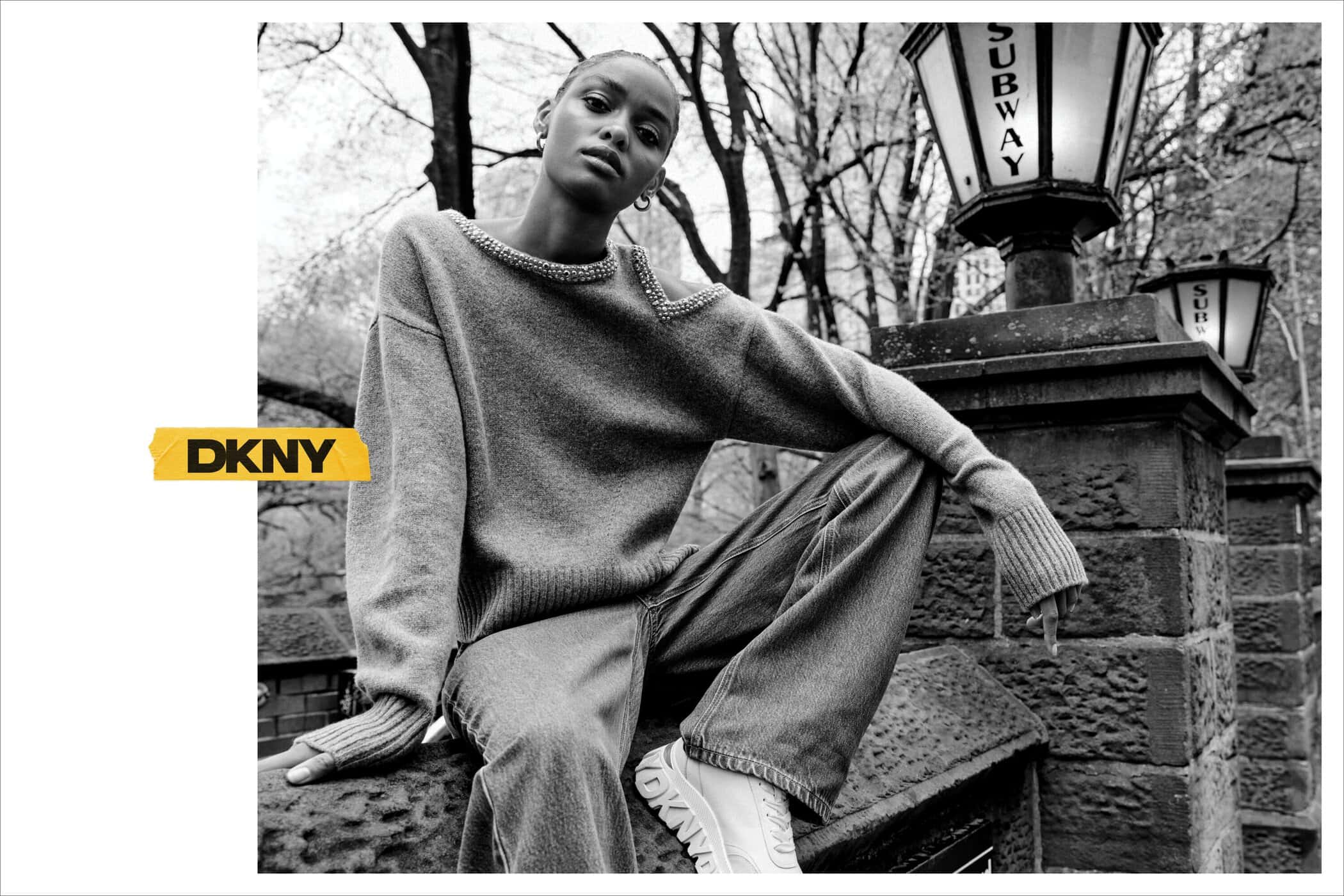 Model wearing a textured knit sweater and loose denim jeans seated outdoors near a subway lamppost in a black and white DKNY ad campaign photograph.