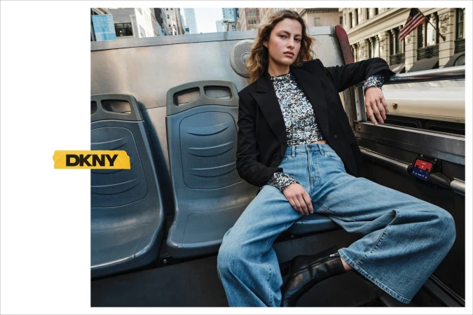 Model seated on the upper deck of a bus wearing a black blazer, sequined top, and blue denim jeans.