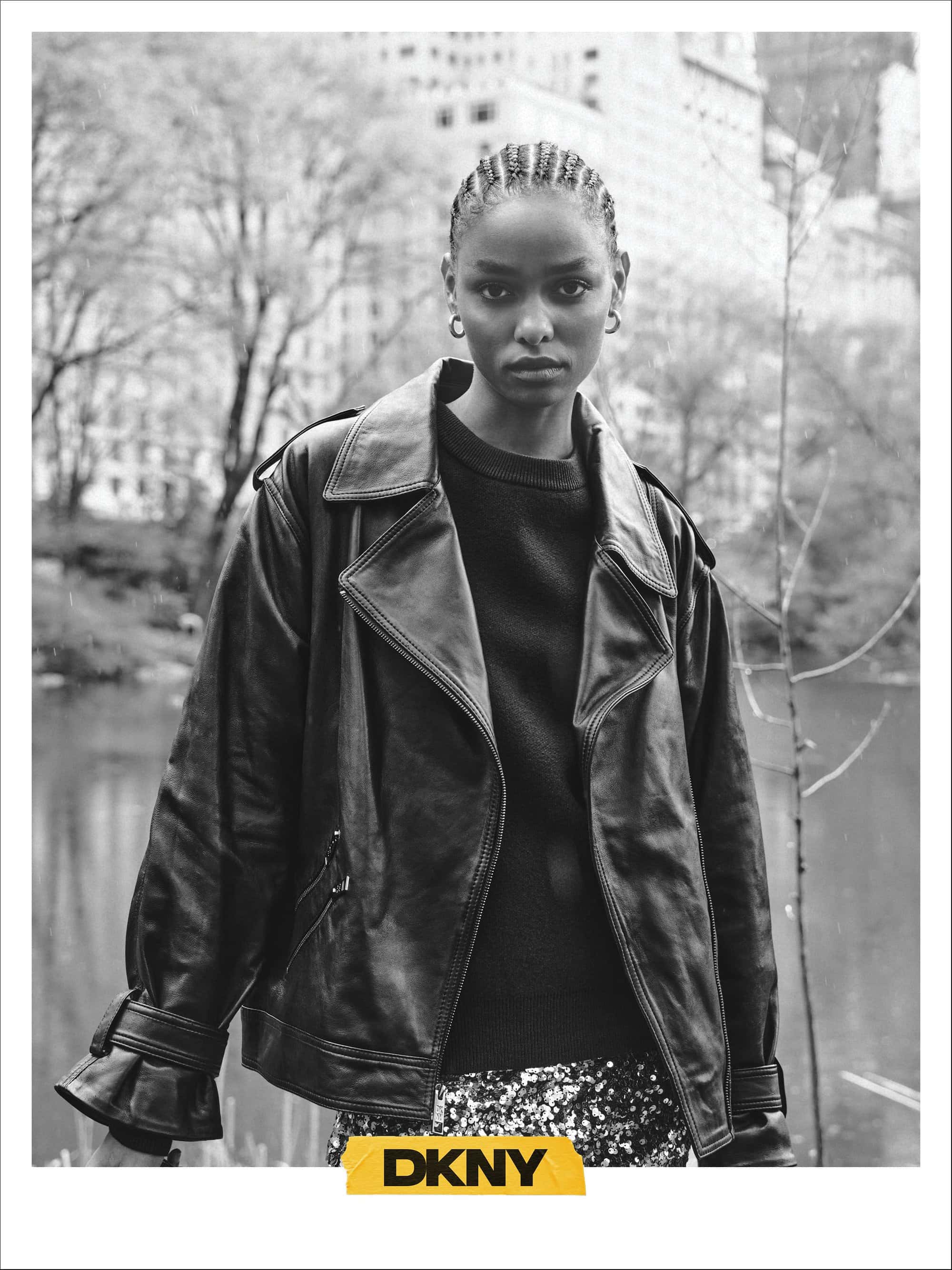 Black and white portrait of a model with braided hair wearing a leather jacket and sweater outdoors near a park pond.