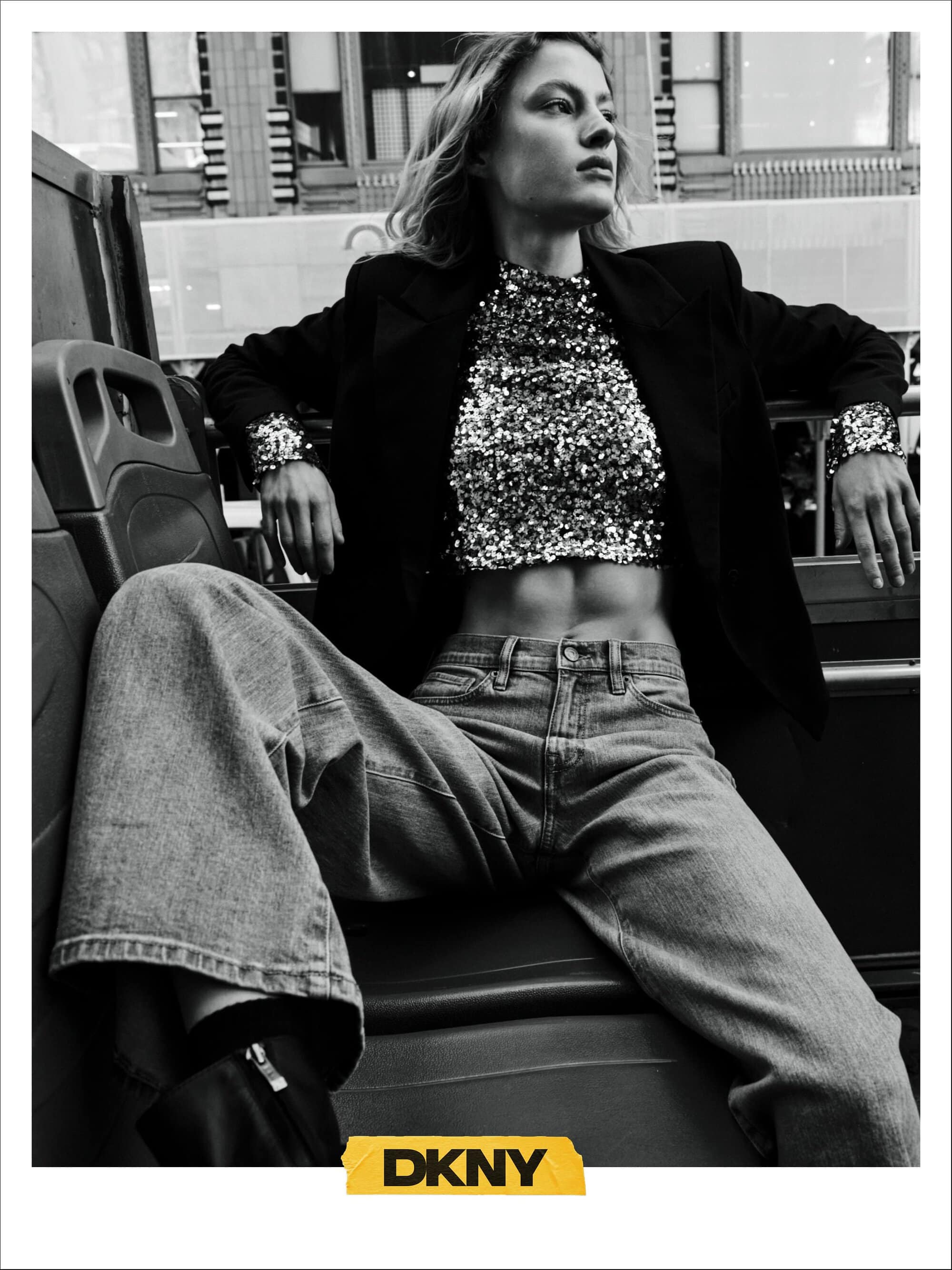 Monochrome fashion campaign image featuring a model seated in a bus wearing a sequined top and denim jeans for DKNY Fall 2024.