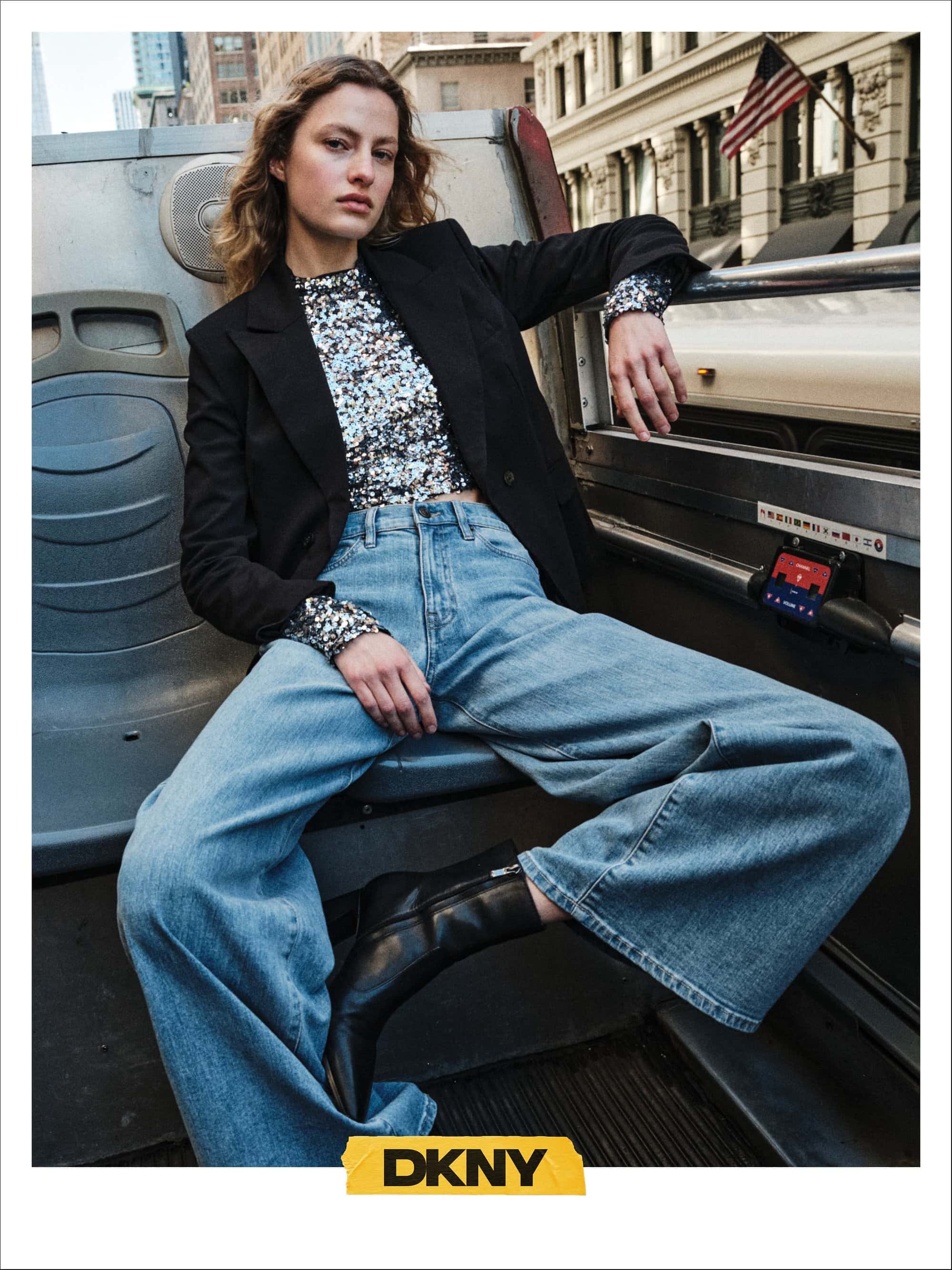 Model wearing black blazer and blue denim jeans seated on top of a city bus
