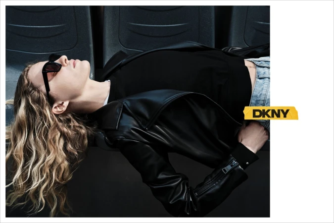 Model with long blonde hair wearing black sunglasses and a black leather jacket reclining against seats for DKNY