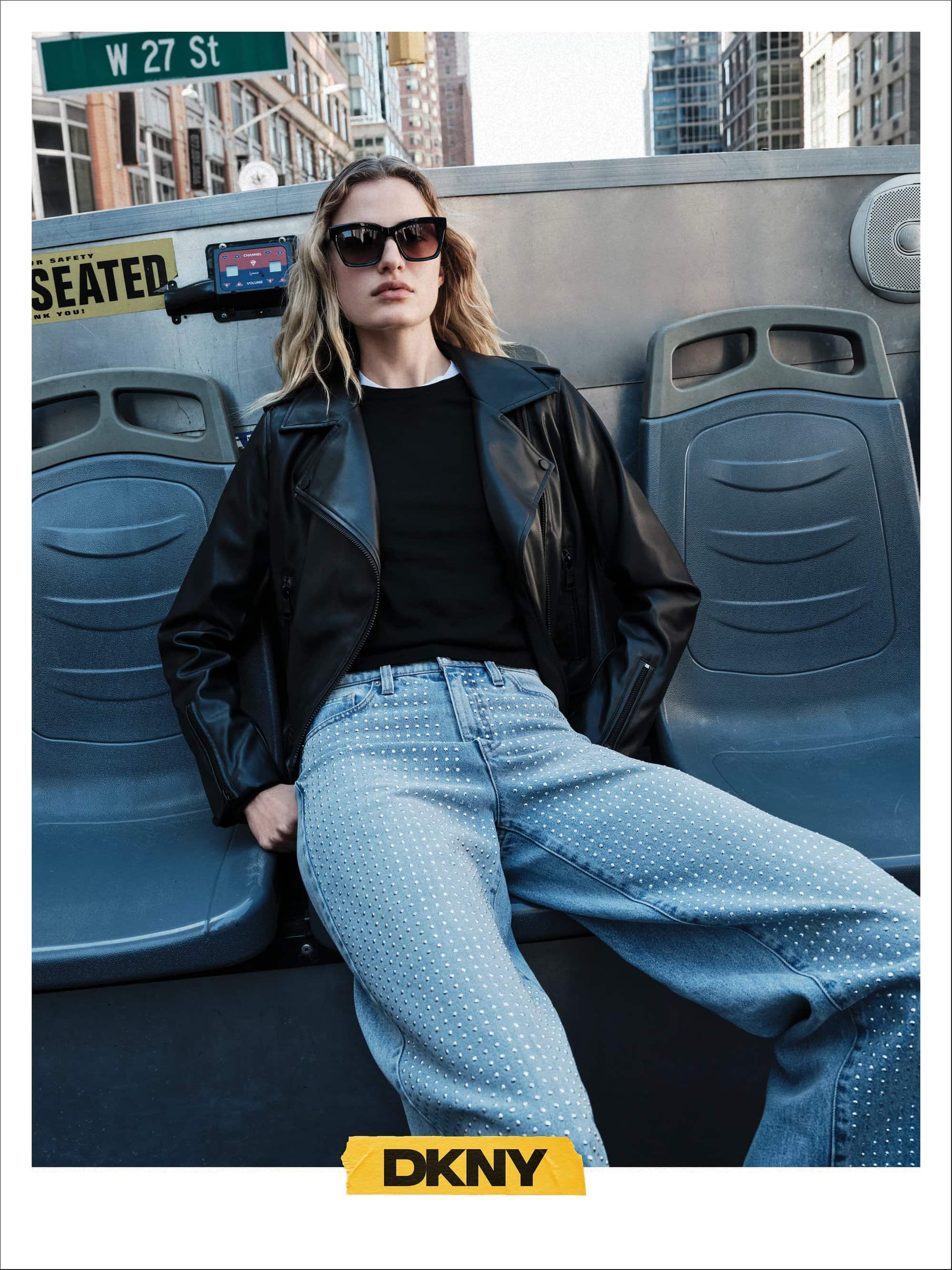 Model wearing dark sunglasses, black jacket, and light denim jeans seated on an upper deck bus in New York