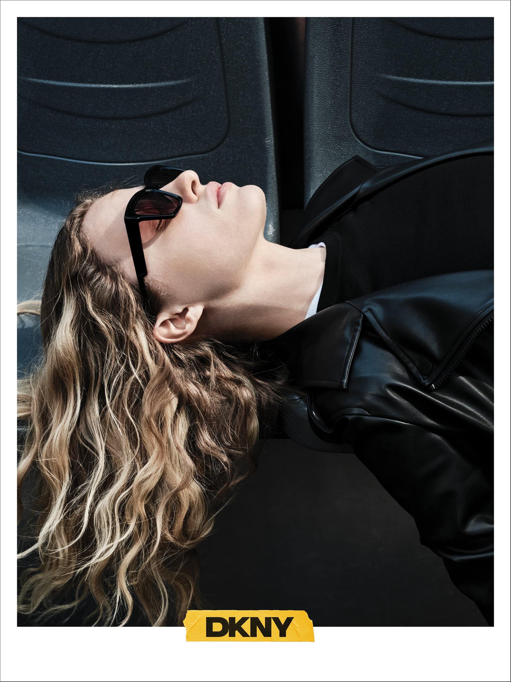 Model with blonde curly hair wearing black sunglasses and a black leather jacket reclining against seats for DKNY campaign.