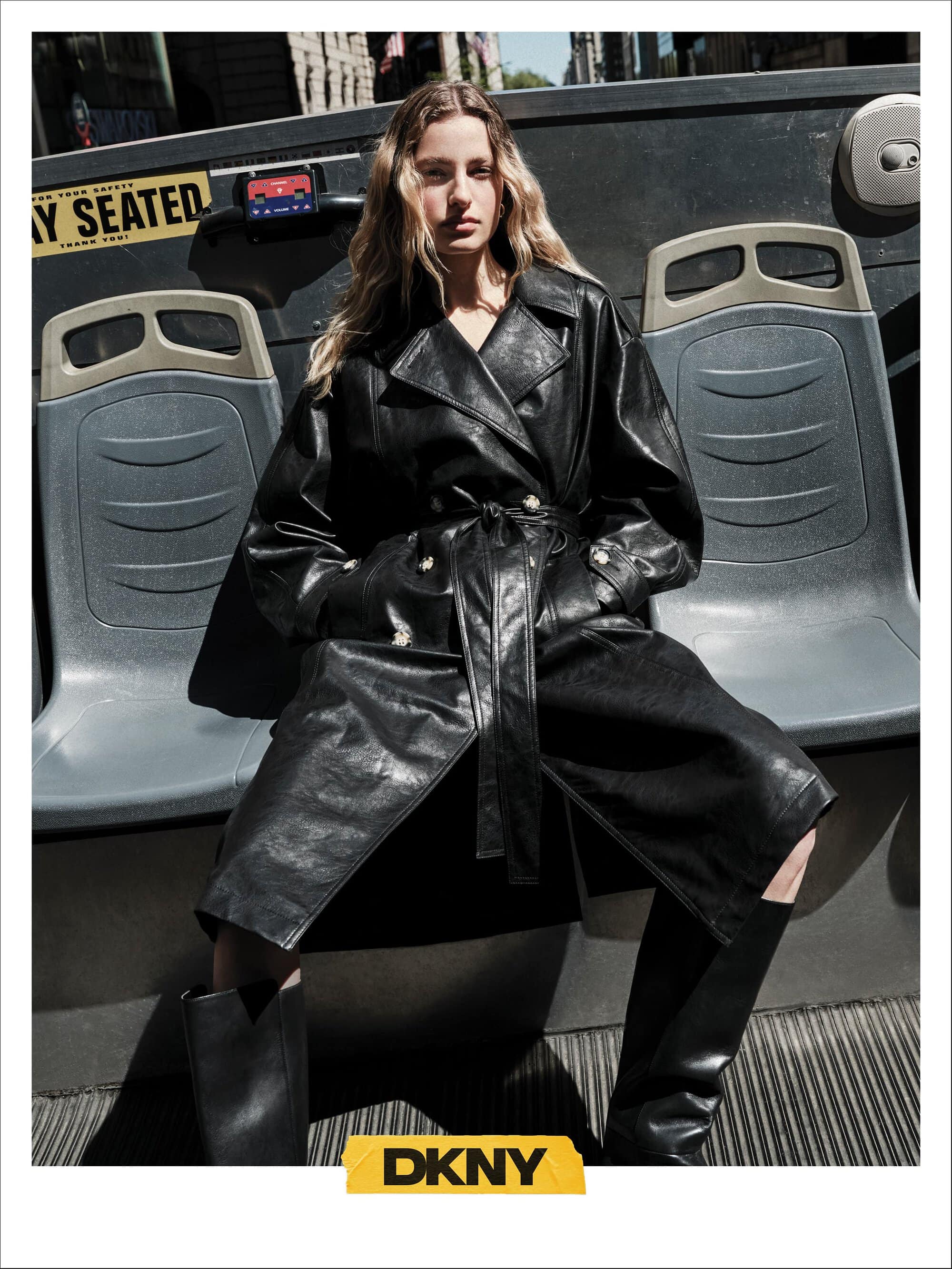 Model wearing a black leather trench coat seated on an upper deck bus seat for DKNY