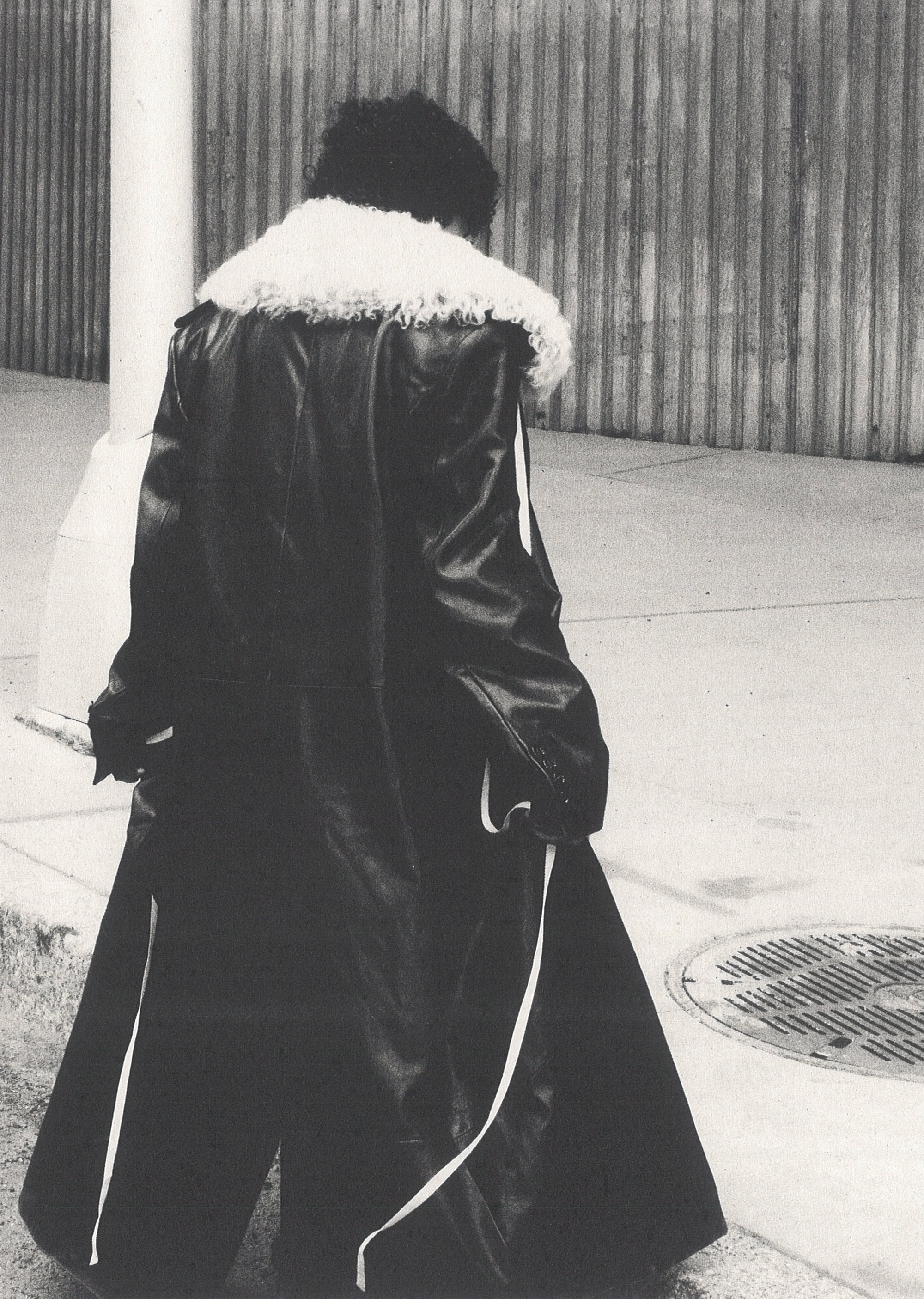Figure from behind walking on a city sidewalk wearing a long black coat with a white fur collar