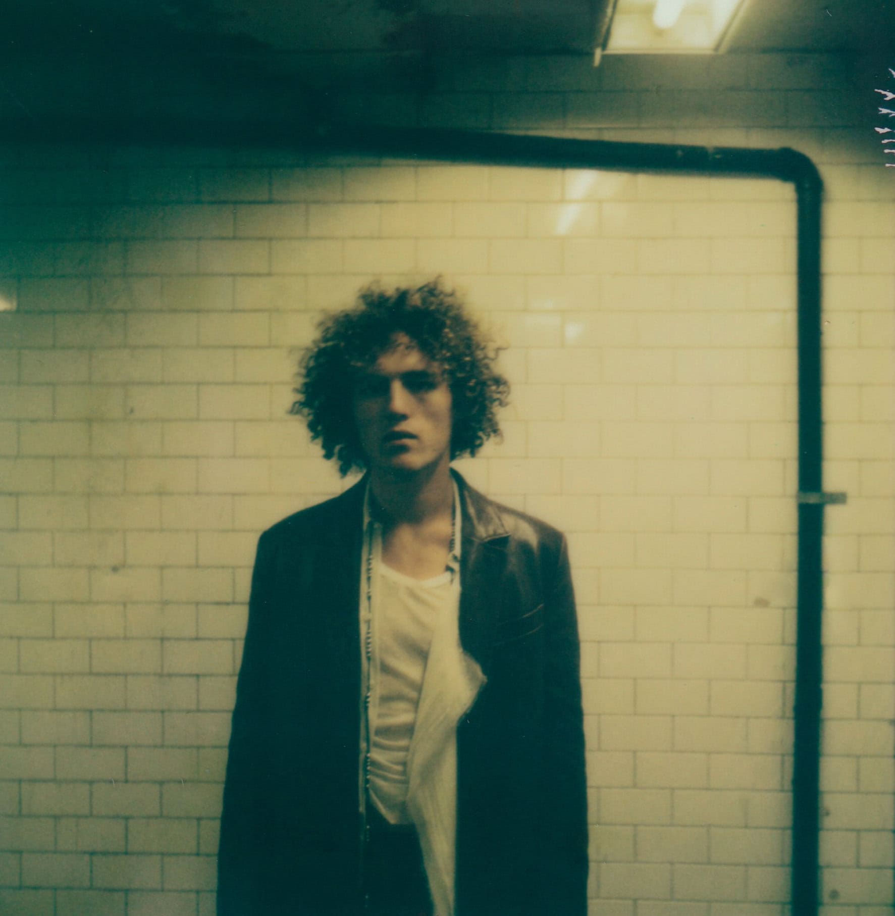 Young man with curly hair wearing a dark jacket over a white top against white tiled wall