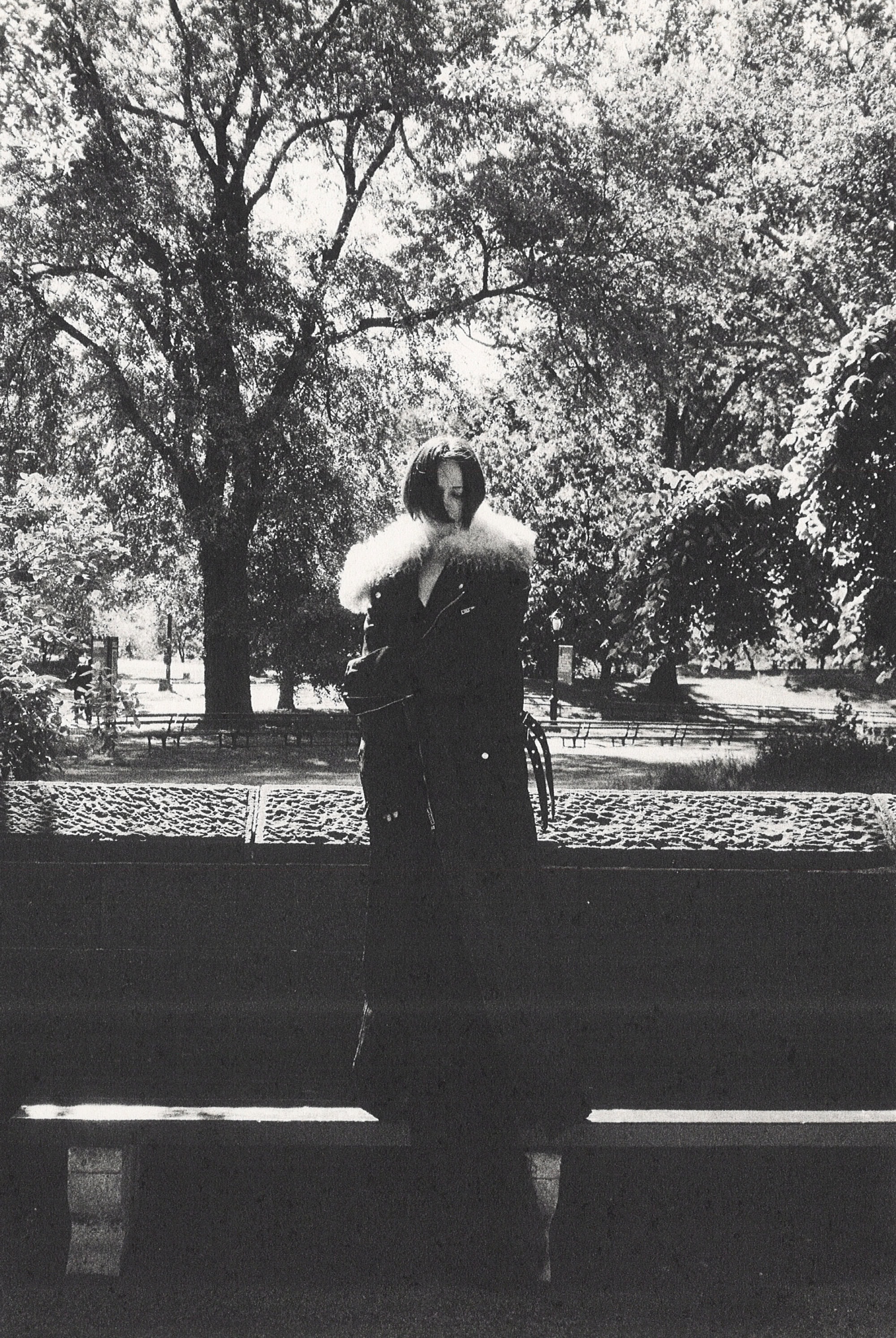 Monochrome fashion campaign image of a model in a dark winter coat with a fur collar standing in a park.