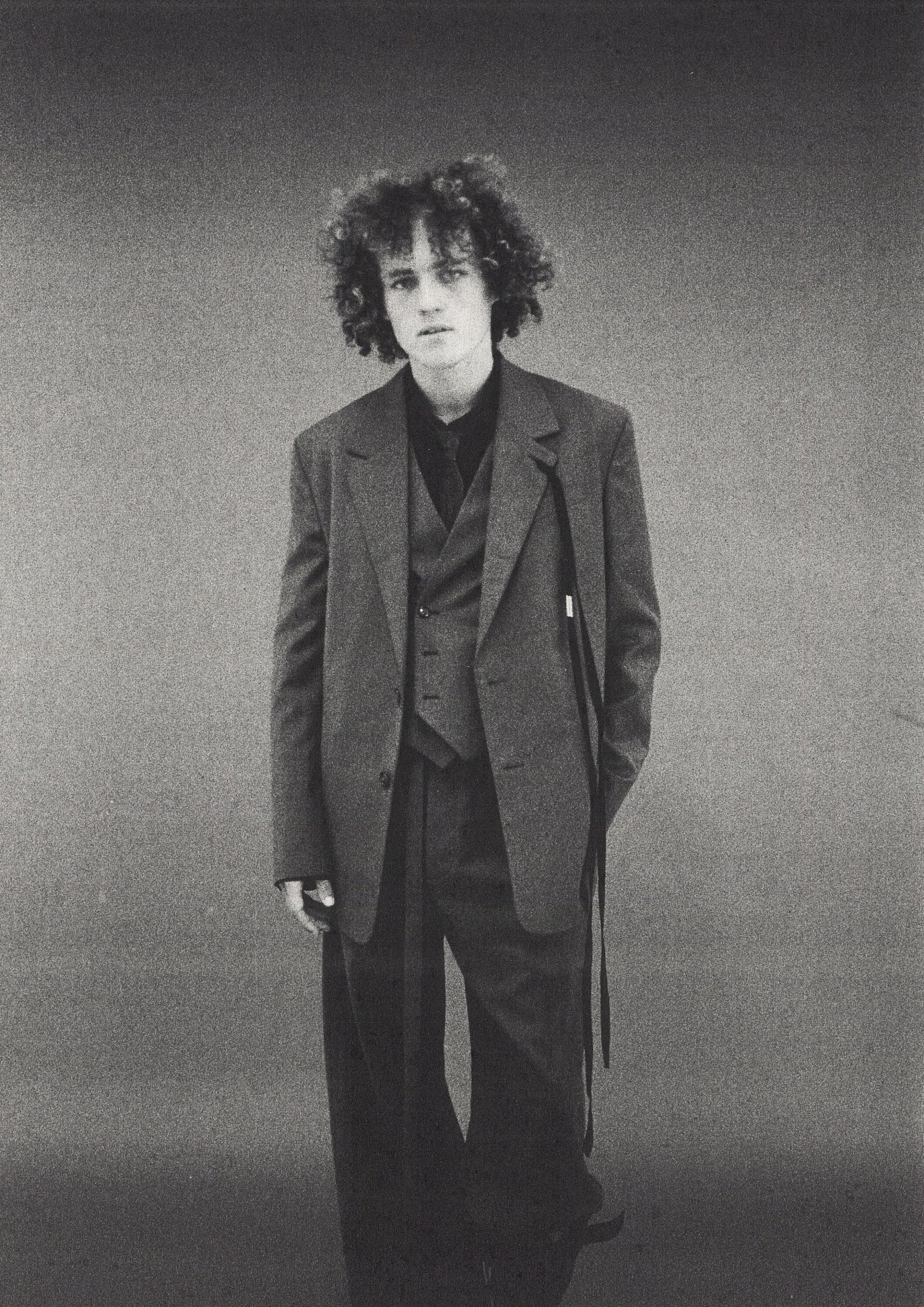 Monochrome portrait of a young man with curly hair wearing an oversized dark suit jacket against a seamless grey studio background.