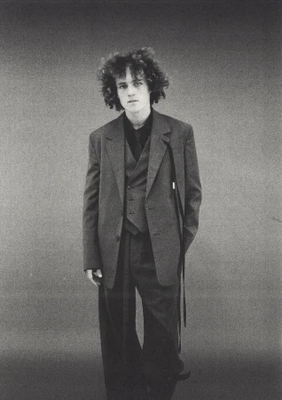 Monochrome portrait of a young man with curly hair wearing an oversized dark suit jacket against a seamless grey studio background.