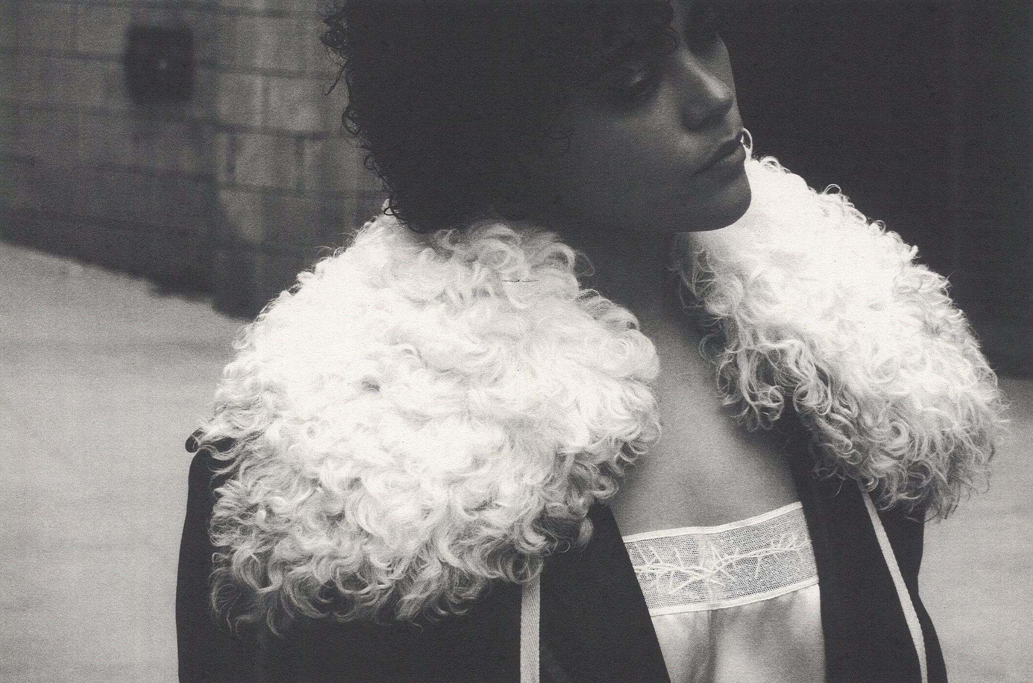 Model wearing dark coat with fluffy white fur collar outdoors against an urban stone wall.