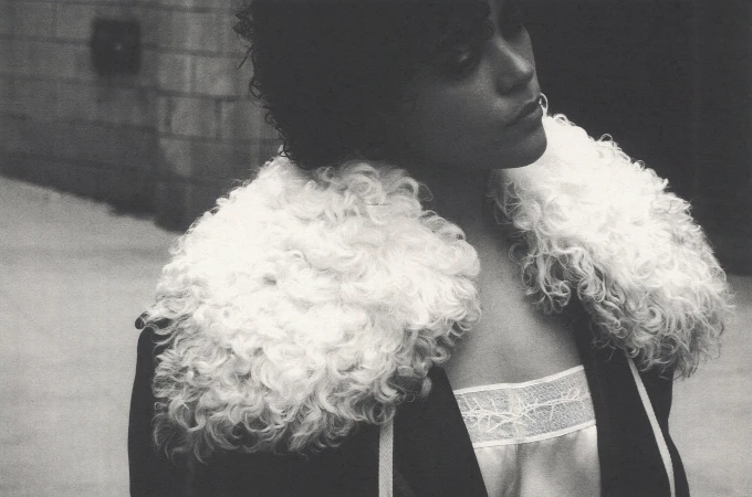 Model wearing dark coat with fluffy white fur collar outdoors against an urban stone wall.