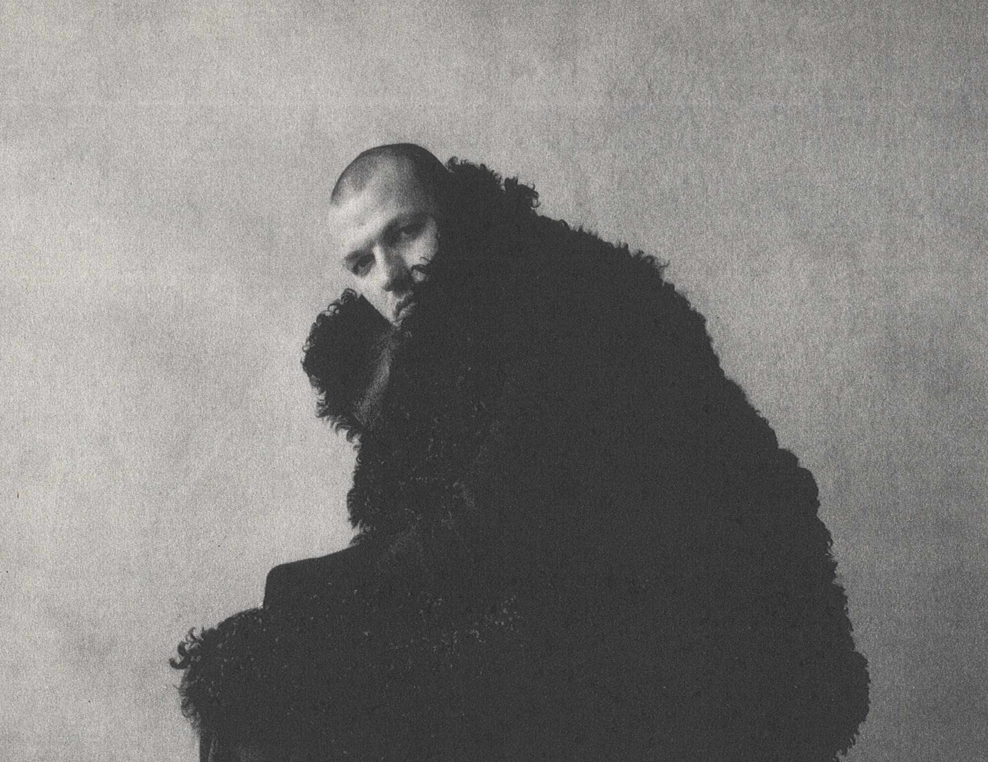 Monochrome portrait of a person in a heavy textured black faux fur coat against a textured backdrop.