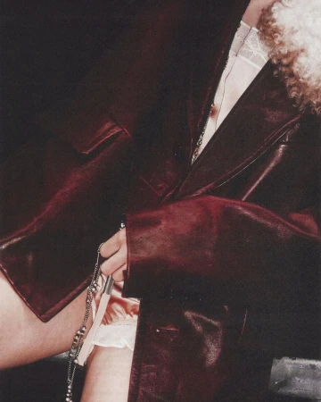 Close-up of a person wearing a dark burgundy leather coat holding a silver chain accessory against a dark background.