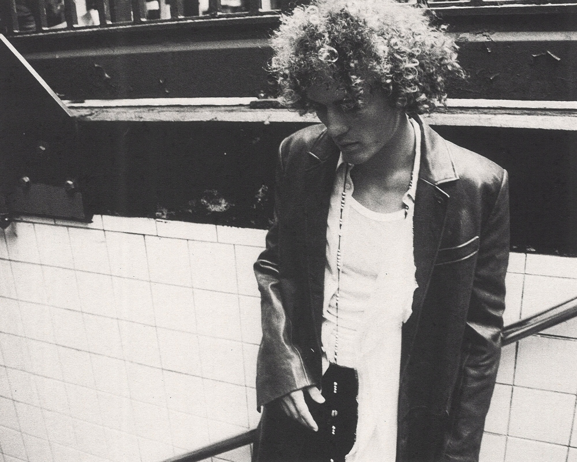 Monochrome portrait of a person with curly hair wearing a leather jacket descending tiled stairs for Ann Demeulemeester