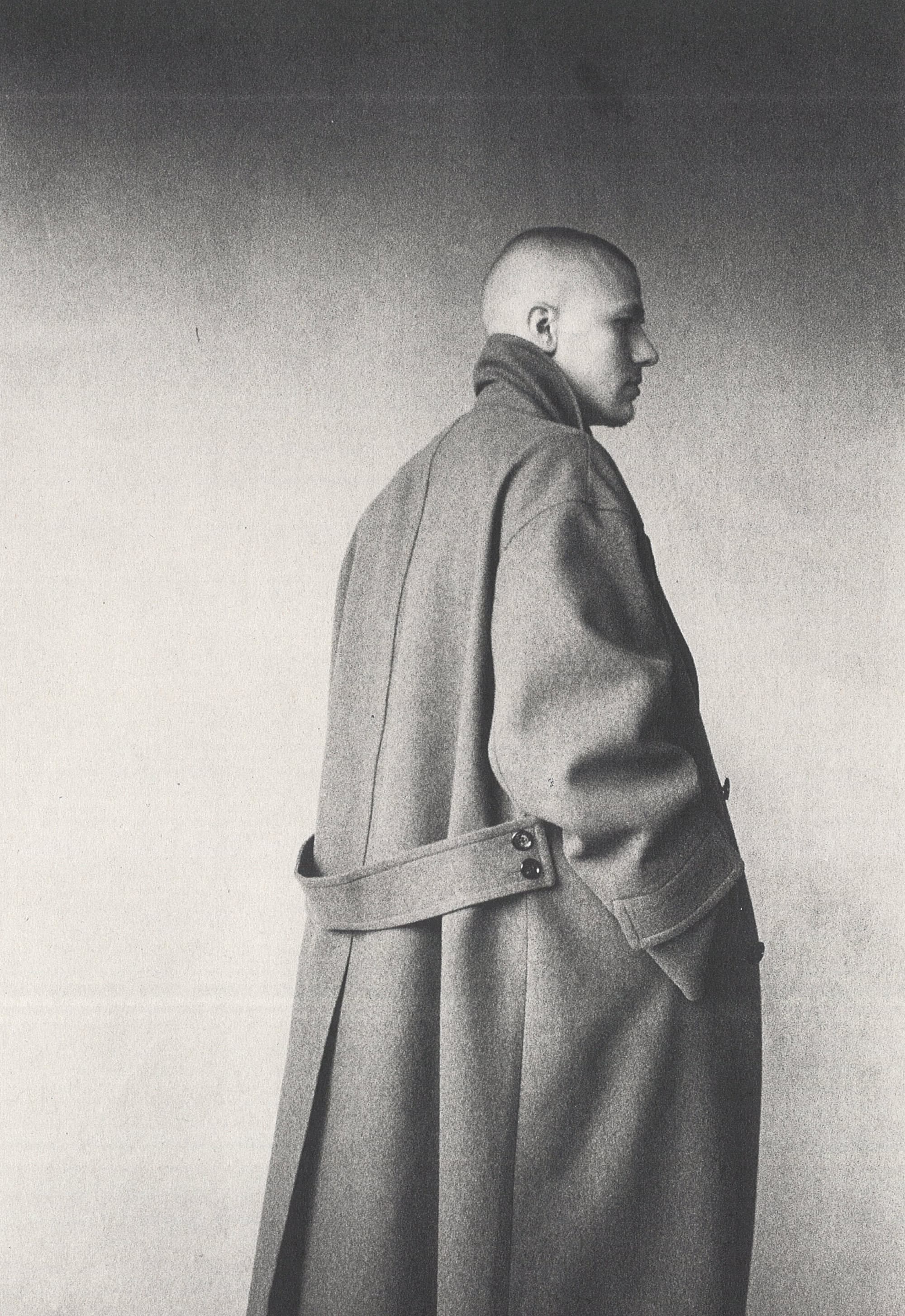 Monochrome portrait of a person in a long grey belted coat standing against a textured studio wall.