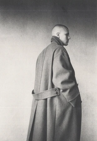 Monochrome portrait of a person in a long grey belted coat standing against a textured studio wall.
