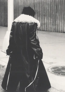 Figure from behind walking on a city sidewalk wearing a long black coat with a white fur collar