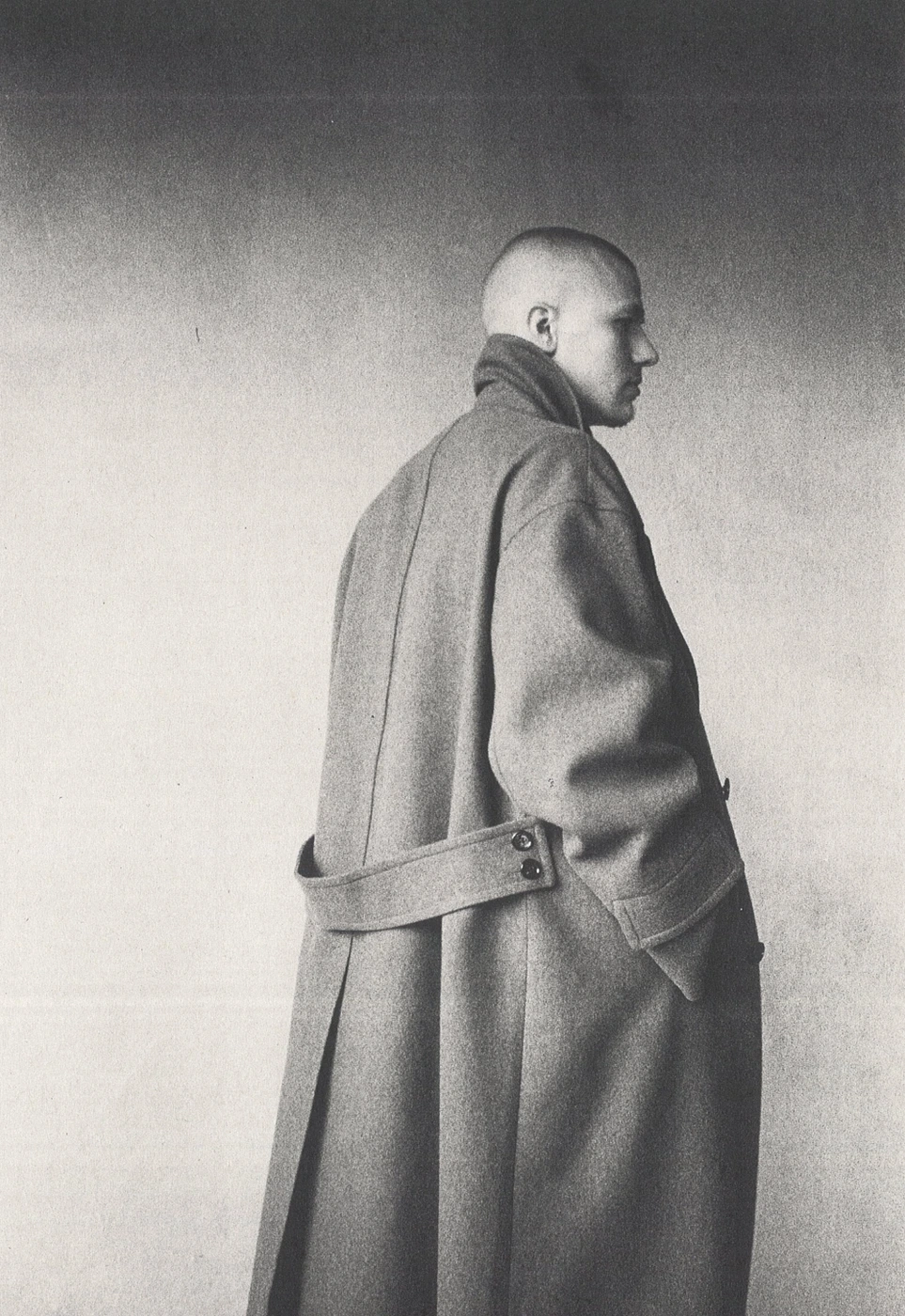 Monochrome portrait of a person in a long grey belted coat standing against a textured studio wall.