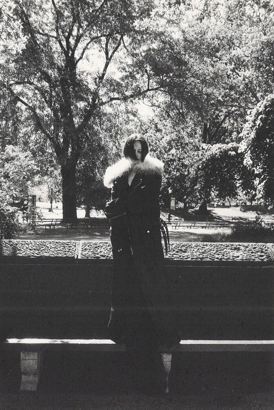 Monochrome fashion campaign image of a model in a dark winter coat with a fur collar standing in a park.