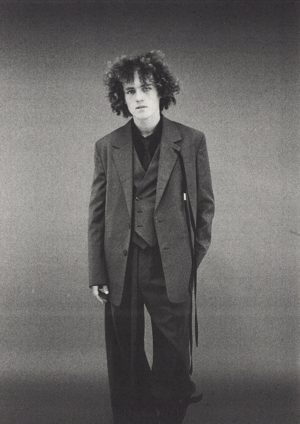 Monochrome portrait of a young man with curly hair wearing an oversized dark suit jacket against a seamless grey studio background.