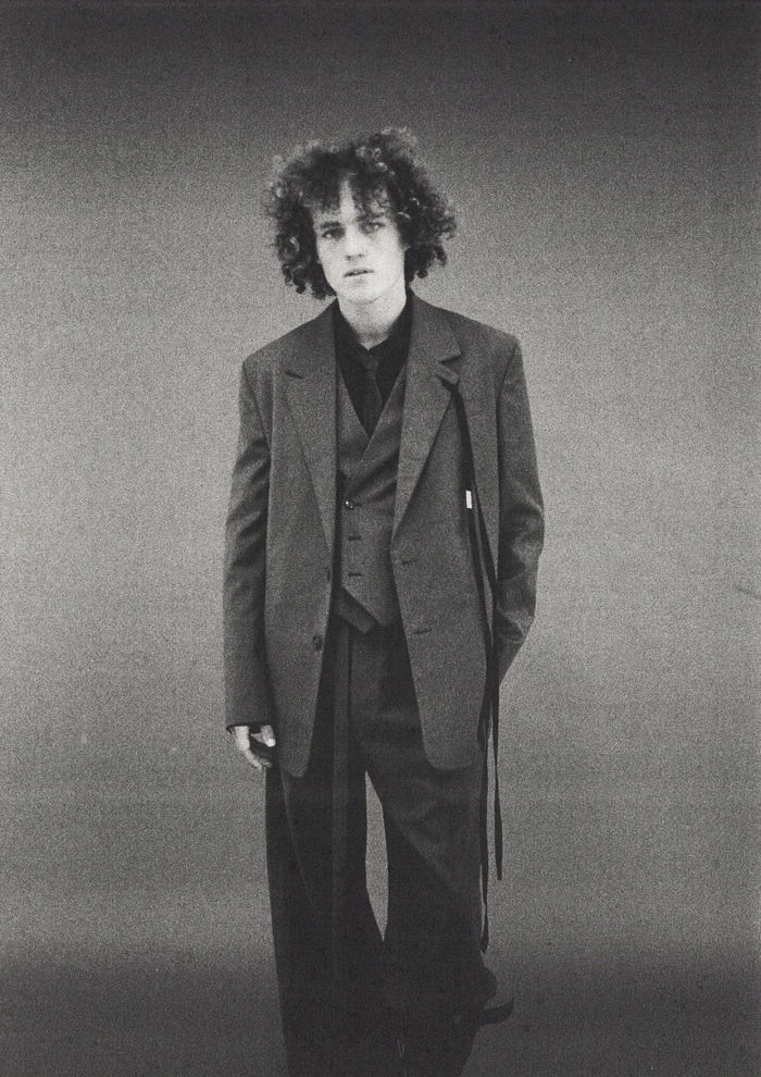 Monochrome portrait of a young man with curly hair wearing an oversized dark suit jacket against a seamless grey studio background.